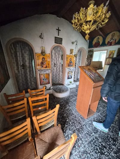 The interior of a small Orthodox chapel in the Municipal Unit of Loutraki - Perachora, Greece, is shown from a low angle. The room features light-colored, textured walls and a dark wooden ceiling. The floor is covered with dark, speckled square tiles. A black Latin cross is mounted on the central wall, positioned above three arched recesses or doorways draped with light-colored, patterned fabric curtains. Multiple religious icons are displayed on the walls, including several framed rectangular icons at eye level and circular icons positioned higher on the right wall. Two brass oil lamps hang from chains in front of icons. A large, ornate golden chandelier with multiple light fixtures is suspended from the ceiling on the right. In the foreground-left, six light-brown wooden chairs with woven rush seats are arranged in rows, facing away from the viewer. To the right, a wooden lectern with an icon displayed on its slanted top stands upright. A person is partially visible on the far right, from the waist down, wearing dark blue jeans and light blue/grey athletic shoes with green accents on the soles. The person is standing, oriented towards the lectern and the icons on the wall.
