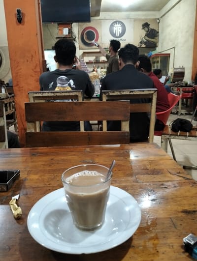 The image captures an indoor scene, likely within a casual coffee shop or cafe, with a focus on a glass of coffee in the foreground and people in the background.
In the immediate foreground, a glass filled with a light brown, milky coffee sits on a white saucer. Small bubbles are visible on the surface of the drink, and a silver spoon rests within the glass. The glass and saucer are placed on a rustic wooden table with a visibly worn, textured surface. To the left of the glass, a black box, possibly a cigarette pack or an ashtray, and a crumpled golden wrapper are visible on the table.
The midground reveals the backs of several patrons seated at wooden tables and chairs. Closest to the camera, the wooden backrests of two chairs frame the scene. Behind these, three men are seated. The man on the left wears a dark long-sleeved shirt featuring a white and yellow graphic of an astronaut on its back. The man in the center is also wearing a dark, possibly black, long-sleeved shirt, facing away from the camera. To his right, another man in a red shirt is partially visible, also seen from behind.
The background shows the interior of the establishment. On the left, an orange-painted pillar or wall segment is visible, with a small black device (possibly a security camera or speaker) mounted on it. A flat-screen television is mounted higher up on this structure. Behind the seated patrons, there is a counter area where a barista is standing. This individual, wearing a dark shirt, is holding an object in their hand and appears to be interacting with the wall decor or another person.
The wall behind the counter is adorned with large graphics. A prominent central logo features a stylized "MR" with a crown above it and the word "Coffee" written below. To the left of this logo is a circular graphic, possibly representing a coffee cup or a target design. To the right, a monochrome illustration depicts a man wearing a fedora hat, playing what appears to be a saxophone or trumpet.
Further into the background on the right, red chairs are visible, suggesting additional seating. The overall lighting is artificial, casting a warm glow on the wooden surfaces and indicating it might be evening or nighttime. The atmosphere appears casual and relaxed, with people enjoying their time in the cafe.