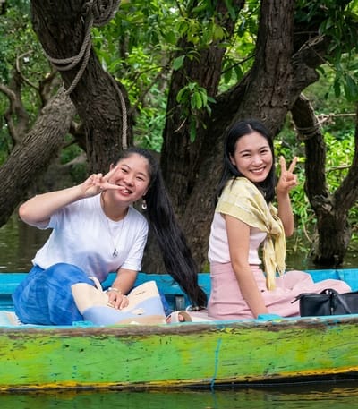 Two young Asian women are seated in a brightly colored boat on a body of water. The woman on the left, with long dark hair pulled back in a ponytail, wears a white t-shirt and a blue skirt. She is smiling and holding up two fingers in a peace sign. The woman on the right wears a white top, a yellow scarf, and a pink skirt. She is also smiling and making a peace sign with her fingers. Behind them, a lush green forest with large trees is visible. The boat appears to be in a natural environment, possibly a lake or river in a forested area. The overall scene conveys a sense of happiness and enjoyment. The location is identified as the city of Soutr Nikom, Cambodia.