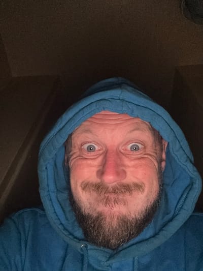 A close-up, low-angle, selfie-style, indoor shot features a Caucasian man with fair skin and blue eyes, wearing a bright blue hooded sweatshirt. His face is framed by the hood, and he has wrinkles around his eyes and forehead, suggesting middle age. He sports a light beard and mustache. His expression is animated, with wide-open eyes and a slight smile, implying a playful or surprised demeanor. The background is dark and indistinct, with a textured appearance that is consistent with an indoor environment. The lighting appears to be artificial and low, casting shadows on the man's face and hood. There is no discernible context of the city of Cheltenham, United Kingdom within the image.