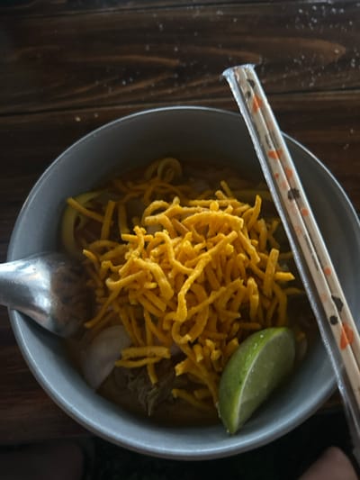 A top-down view shows a grey-blue ceramic bowl containing a serving of Khao Soi. The bowl holds an orange-yellow curry broth with submerged soft egg noodles, pieces of meat, and sliced white onions or shallots. A prominent layer of crispy, fried yellow egg noodles is piled on top. A wedge of green lime rests on the right edge of the bowl. A metallic spoon is partially submerged on the left side of the bowl. A pair of white chopsticks, featuring orange and black patterns, are enclosed in a clear plastic sleeve and rest diagonally across the right side of the bowl. The bowl is situated on a dark brown wooden surface, exhibiting visible grain, with faint white specks scattered across it. The scene is located in the city of Ban Paem Bok, Thailand.