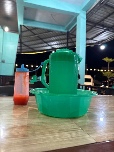 In Grong-Grong, Indonesia, a green plastic pitcher sits inside a matching green plastic basin on a wooden table. A clear plastic squeeze bottle with a blue lid, filled with a red liquid, is positioned to the left of the pitcher. The background reveals an outdoor seating area with a metal roof supported by teal-colored pillars. String lights are visible in the distance, illuminating the night sky. A white van and a small tree are also present in the background.
