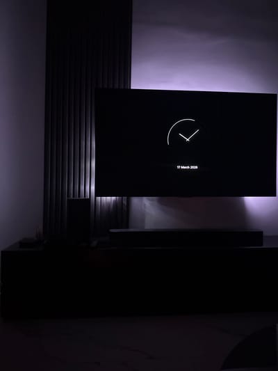 The image depicts an indoor scene in Latsia, Cyprus, characterized by low light with a predominant purple-pink ambient illumination. A large, dark-screened television is centrally positioned, displaying a white minimalist digital clock face with hands and the text "17 March 2026." Directly beneath the television is a dark soundbar. Below the soundbar, a large, dark, rectangular media console or cabinet spans the width of the lower frame. To the left of the television, a vertical wall section is covered with dark, ridged or slatted panels, extending from the bottom of the television upwards. The wall behind the television and slatted panel is light-colored and glows with the purple-pink light, suggesting a hidden light source. The floor in the foreground is a light-colored surface, possibly polished stone or marble, exhibiting subtle patterns. No individuals, actions, or direct interactions are depicted.