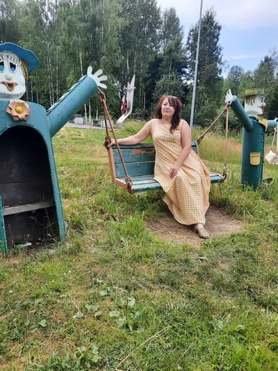 In Зеленогорский, Russia, a woman with dark hair, wearing a light yellow checkered dress and olive green sandals, sits on a weathered teal-colored swing. The swing is suspended by thick ropes connected to a tall, dark pole. To her left, a large, teal-colored scarecrow-like figure with a blue hat, painted face, and yellow flower accent stands beside the swing. To her right, another similar scarecrow figure, also teal with a hat and painted face, is visible. The figures appear to be part of a playground or decorative installation. The ground is covered in green grass with some dry patches and scattered small white flowers. In the background, a dense forest of green trees, primarily birches, creates a natural backdrop under a bright, partly cloudy sky.