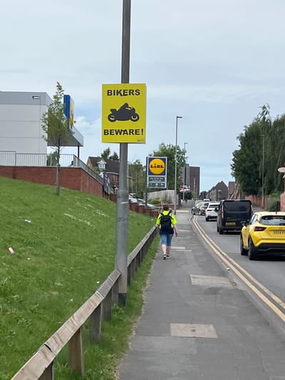 The image depicts a street scene in Stoke-on-Trent, United Kingdom. On the left side of the frame, a bright yellow sign with black text and a silhouette of a motorcycle reads "BIKERS BEWARE!". Adjacent to the sign is a modern, white-fronted building, likely a commercial establishment, with a red brick wall and a grassy slope in the foreground. A person wearing a neon yellow vest and dark shorts is walking away from the camera along the sidewalk, which runs alongside the road. Several cars, including a yellow SUV and a black van, are present on the road. The sky is overcast. In the background, residential buildings and trees are visible. The overall scene suggests an urban or suburban environment.