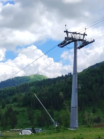 The image depicts a ski lift in Bakuriani, Georgia. A tall, grey metal tower supports the ski lift's machinery and cables, which extend diagonally across the frame against a backdrop of a lush, green mountainside. The mountain is covered with dense forest and a bright green meadow at its base. In the distance, a rounded hill is visible, topped with communication towers. The sky is a mix of bright blue and fluffy white clouds. At the base of the ski lift, a small building with graffiti and a row of colorful ski lift chairs are present.