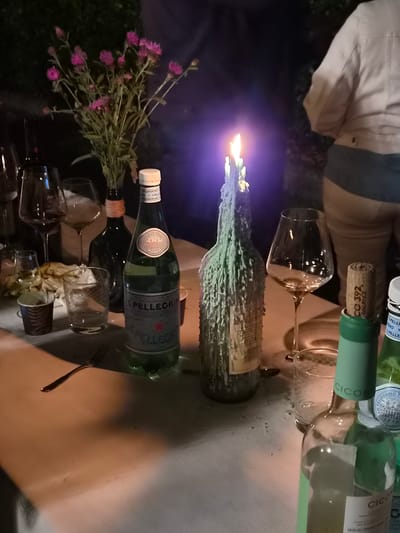 The image depicts a dimly lit outdoor scene, likely a dinner party, set on a table covered with a white tablecloth. The table is laden with various items including several wine glasses, two bottles of San Pellegrino mineral water, a decorative bottle with melted wax dripping down its sides and a lit candle, a small vase with pink flowers, and a single fork. In the background, a person wearing a light-colored jacket and dark pants is partially visible, suggesting they are attending the gathering. The overall ambiance is relaxed and festive, with the flickering candlelight adding a warm glow. The location is identified as Bettembourg, Luxembourg.