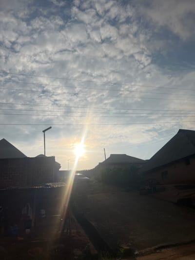 This image captures a street scene in Gembu, Nigeria, bathed in the warm glow of the sun. The sun is positioned high in the sky, casting bright rays that create a lens flare effect. The sky is filled with fluffy, white clouds against a light blue backdrop, suggesting fair weather.
The foreground and right side of the image are dominated by the shadows and shapes of buildings, with roofs and walls visible. Some of these structures appear to be made of brick, while others have metal roofing. A few sparse green plants are visible near the base of some buildings, indicating some vegetation despite the urban setting.
Several power lines are strung horizontally across the image, a common sight in many urban and semi-urban environments. A street lamp is also visible, its arm extending outwards. The overall impression is that of a residential or mixed-use area.
The low angle of the sun and the long shadows suggest it is either early morning or late afternoon. There are no people clearly visible in the image, but the presence of the buildings and power lines implies human habitation and infrastructure. The scene evokes a sense of calm and a typical day in a Nigerian town. No visible text is present.