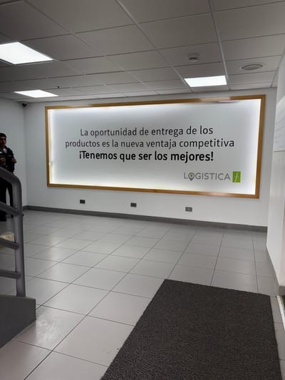 The image shows an interior view of a building, likely an office or commercial space, with a large illuminated sign on the wall. The sign contains text in Spanish and a logo. The text reads: "La oportunidad de entrega de los productos es la nueva ventaja competitiva ¡Tenemos que ser los mejores! LOGISTICA f". Below the sign, two electrical outlets are visible on the wall. The floor is tiled with large white square tiles. To the left, a staircase with a metal railing is partially visible, and a security guard in uniform is standing near the stairs. The ceiling is made of modular tiles with fluorescent lighting panels. A dark gray doormat is placed on the floor in the lower right portion of the image. The overall environment suggests a professional and functional space. The image does not contain any explicit information placing it in Villa El Salvador, Peru, but the text on the sign indicates a business related to logistics.