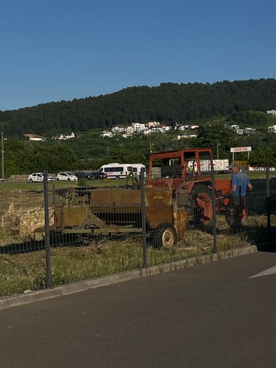 A man wearing a blue t-shirt and a brown cap stands behind a metal fence, next to an orange tractor with a trailer attached. The tractor and trailer appear old and rusty. The man is facing towards the right of the frame and looking forward. Behind the tractor, there are several vehicles, including a white bus and cars. In the background, there is a wooded hill with white houses scattered across it. A sign with the word "ONCOS" is visible on the right side of the frame. The scene appears to be located in a semi-rural or suburban area.