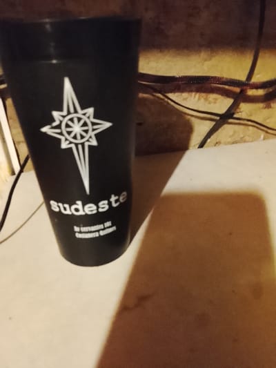 The image depicts an indoor scene in the city of Bernal Este, Argentina, featuring a black insulated cup resting on a light-colored surface against a textured background.
The central subject is a black, tall, cylindrical cup or tumbler positioned in the left-central portion of the frame. It displays white printed graphics and text. A prominent stylized compass rose or star symbol with eight points is located on the upper half of the cup. Below this symbol, the word "sudeste" (Spanish for "southeast") is printed in white, lowercase letters. Further below, in smaller white text, are the lines "bv. Cervantes 101" and "Costanera Quilmes," suggesting a specific address and a location relevant to the wider Bernal/Quilmes area.
The cup rests on a flat, light-beige or off-white surface, possibly a table or counter, which occupies the lower and right-hand portions of the image. This surface shows minor blemishes or dust. An elongated, dark shadow, likely cast by the cup from a light source to the left, stretches across this surface towards the right and bottom of the frame.
The background behind the cup and surface is a darker, textured wall, appearing to be a reddish-brown or dark wood tone. Multiple dark electrical wires or cables are visible against this background, particularly in the upper right quadrant, some appearing bundled or intertwined. The overall lighting is dim, creating distinct shadows and moderate contrast. No people or active interactions are present in the scene.