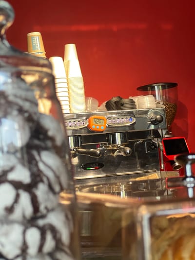 This media file shows a close-up, rotated view of an espresso machine in what appears to be a cafe or food service establishment. The machine is predominantly made of stainless steel with some red accents. On the left side, a clear hopper filled with coffee beans sits atop the machine. Below the hopper, stacks of white disposable paper cups are visible, some within a dispenser.

The central part of the machine features a control panel with several buttons and a small orange digital display showing "42.0". Above this, a row of circular buttons with illuminated blue indicators is present. A silver lever, likely for dispensing espresso, is positioned to the right of the control panel. To the far right, a black oval indicator with green, yellow, and red lights is visible. The background is a solid, textured red wall.

In the foreground, another stack of disposable cups can be seen, with one cup showing some text that is difficult to read clearly but appears to be a logo or pricing information. The overall scene is well-lit, suggesting an indoor setting, possibly during business hours. There are no people visible in the frame. The focus is on the intricate details of the espresso machine and its associated supplies, highlighting the process of coffee preparation.