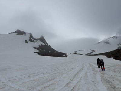 The image depicts a snowy, mountainous landscape under a cloudy sky, with two individuals walking away from the camera on a snow-covered path. The terrain is predominantly white with visible tracks in the snow, suggesting recent passage. Darker rocky outcrops are visible on the slopes of the mountains in the background, which are partially obscured by fog. The individuals are dressed in outdoor clothing suitable for cold weather. The person in front is wearing a dark jacket and pants, and the person behind is wearing a grey hooded jacket and dark pants, with bright orange and blue boots. The scene is devoid of any urban elements, indicating a remote, natural environment. The location is identified as Petropavlovsk-Kamchatskiy, Russia, but the visual information in the image itself does not provide any direct clues to confirm this specific urban locality.