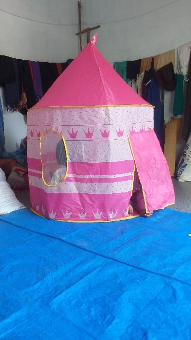 An indoor scene in the city of Cot Madi, Indonesia, depicts a pink, conical play tent centrally positioned on a blue tarp covering the floor. The tent's lower section is light pink with darker pink horizontal stripes, white polka dots, and repetitive pink crown motifs. A person's head and upper torso, wearing a light-colored striped garment, are partially visible through an oval-shaped, transparent window with a yellow border on the tent's left side. Two bare feet are visible at the base of the tent's right-side entrance, which is open. The background features a light-colored wall from which various fabric items, including scarves and garments in dark blue, purple, red, brown, green, and white, are suspended. A wooden cross-like structure is affixed to the wall in the upper center. A large white sack is partially visible on the far right. The immediate environment suggests a functional living or activity space.