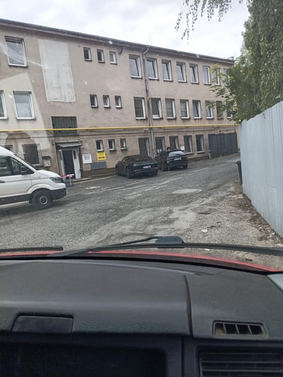 The image shows a view from inside a vehicle looking out at a building and parked cars in what appears to be an industrial or commercial area in Svémyslice, Czechia. A light-colored van is parked on the left, and two dark-colored cars are parked further down on the right. The building behind them is a multi-story structure with numerous windows. The ground is paved with a rough, textured surface, and there is a tall, corrugated metal fence on the right side of the frame. The sky is overcast. The interior of the vehicle, including the dashboard and windshield wipers, is visible in the foreground.