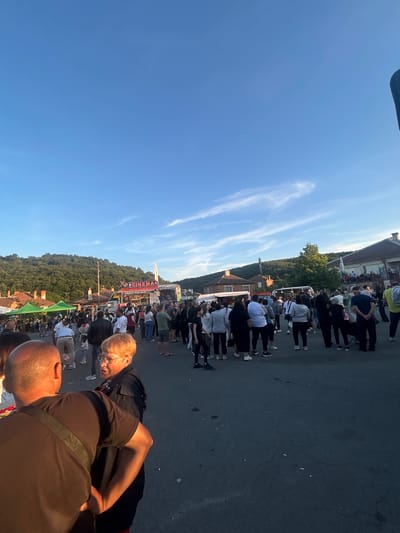 In the city of Gabar, Bulgaria, a public gathering takes place on a paved street. A clear blue sky with wispy clouds stretches above, casting a warm, late afternoon light. The scene is populated by a diverse crowd of people, many of whom are casually dressed, suggesting an outdoor event or festival.
Prominently featured is a large banner with the words "3D CINEMAS" and "ATTRACTION VIRTUAL REALITY," indicating a form of entertainment is present. Tents, possibly for vendors or attractions, are set up along one side of the street, with people milling around them. In the background, traditional-looking buildings with red-tiled roofs are visible, nestled against a backdrop of lush green hills.
Two individuals in the foreground are engaged in conversation. One man, viewed from the back, wears a dark t-shirt and has a bag slung across his body. He appears to be gesturing with his right hand. Facing him is a woman with short, auburn hair and glasses, wearing a dark jacket. Their interaction suggests a casual exchange within the bustling environment. The overall impression is of a lively community event unfolding in a scenic, rural Bulgarian setting.