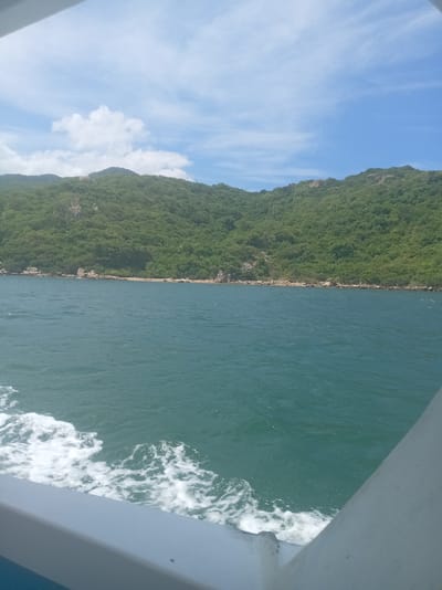 The image captures a scenic view from a boat sailing on the turquoise waters of Vinh Hai, Vietnam. The lush green, tree-covered hills of a coastal mountain range dominate the background, with rocky outcrops visible along the shoreline. The sea is slightly choppy, creating white foam in the foreground as the boat moves forward. The sky above is a clear blue with scattered white clouds. The perspective suggests the viewer is on the deck of a vessel, with a portion of the boat's railing visible in the lower right and lower left corners of the frame. The scene is natural and serene, showcasing the coastal landscape of Vinh Hai.