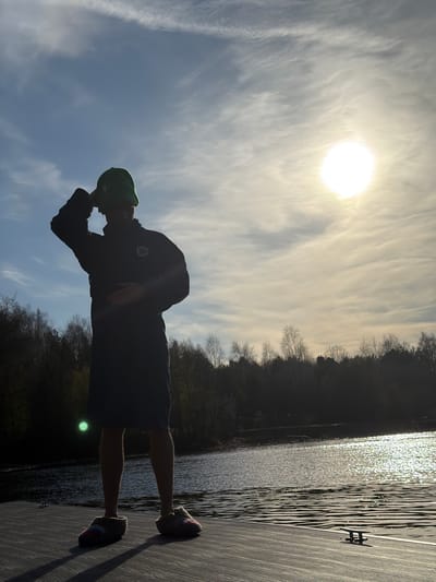 The image, set in Shchelkovo, Russia, portrays a person standing on a wooden dock by a body of water, strongly backlit by the sun. The individual is rendered in silhouette, facing towards the brilliant sun positioned in the upper left of the frame. One hand is raised towards their face, perhaps shielding their eyes from the glare or resting in a contemplative pose. The person is dressed in a dark jacket or sweatshirt, dark shorts, a bright green cap, and fuzzy slippers adorned with colorful bands of red, white, and blue/purple.

The sun is a dominant, overexposed orb in the sky, casting bright light and illuminating delicate, streaky cirrus clouds across the blue expanse. Along the distant bank of the water, a dense line of trees, mostly bare of leaves, forms a dark, silhouetted forest. The water in the mid-ground, likely a lake or river, glistens with numerous sparkling reflections from the sun. The foreground is occupied by the dark, textured surface of the wooden or composite dock, on which the person stands. Long shadows cast by the person and metal cleats on the dock suggest either early morning or late afternoon. The scene conveys a bright, clear day with a contemplative atmosphere. No visible text is present.