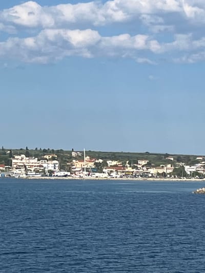The image shows a coastal town in the Municipal Unit of Kastro - Kyllini, Greece. In the foreground, there is a body of blue water with small ripples. In the middle ground, a line of buildings with white and orange facades runs along the coastline, interspersed with green trees. Several boats, including sailboats and smaller motorboats, are docked or moored along the waterfront. The town is built on a gentle slope leading up to a hill covered in green vegetation and scattered houses. The background features a clear blue sky with scattered white clouds. The overall scene depicts a tranquil Mediterranean coastal settlement.