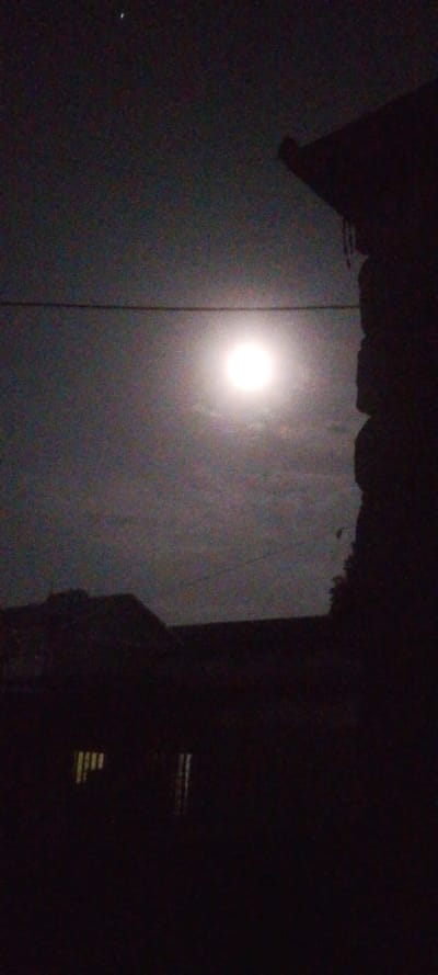 The image presents a night scene with the moon shining brightly in the sky. Silhouettes of buildings and a utility wire are visible in the foreground and mid-ground, obscuring some of the sky. The right edge features a dark structure, likely part of a building. Overall, the image has a dark and somewhat grainy quality, emphasizing the stark contrast between the bright moon and the surrounding darkness.
