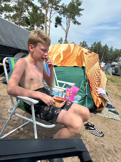 A young boy with blond hair is sitting in a striped camping chair, facing right. He is shirtless and wearing black shorts with a pattern of motorcycles and the German phrase "Ein Moment". He is eating from a bag of chips held in his right hand, with his left hand near his mouth. The boy has a relaxed expression.
The scene is a campsite with tents and trees in the background. A green tent is visible behind the boy, partially covered by a patterned blanket with a tiger-like stripe design. There are other camping items scattered around, including a pair of black and white striped socks on the ground and some dark blue flip-flops. The ground is covered in dirt and dry grass.
The sky is blue with some wispy clouds, suggesting a sunny day. The lighting indicates it is daytime, likely afternoon. The presence of camping gear and the outdoor setting place the scene at a campsite. The overall atmosphere appears to be casual and relaxed, consistent with a camping trip. The text "Ein Moment" is visible on the boy's shorts.