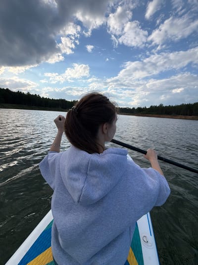 A person in a gray hoodie is seen from above on a stand-up paddleboard on a body of water. They are paddling with a black paddle. The water reflects the sky, which is filled with clouds. In the background is a forest. It is not possible to identify a specific location in Selychka, Russia, from the image alone.