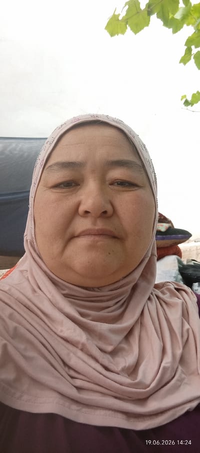 A medium shot selfie features a woman wearing a light pink hijab with beaded trim, covering her head and neck. She is looking directly at the camera with a neutral expression. The background is blurred, showing hints of a dark tent or fabric on the left and stacked objects including blankets or pillows on the right. Green leaves are visible in the upper right corner of the frame, suggesting an outdoor setting. The overall lighting is bright, and the timestamp "19.06.2026 14:24" is present in the lower right corner. The image appears to have been taken in Чоң-Гара, Kyrgyzstan.