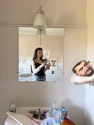 A rotated medium shot depicts a bathroom scene in the Town of Red Hook, United States of America. A mirror reflects a young woman holding a phone, presumably taking a selfie. The mirror has a sign at the bottom warning against pouring solvents or similar materials down the drain. The environment includes a light fixture on the left, a headshot portrait taped above the mirror, and toiletries, including a can of Pola Seltzer, arranged on the right. A water bottle sits near the sink.