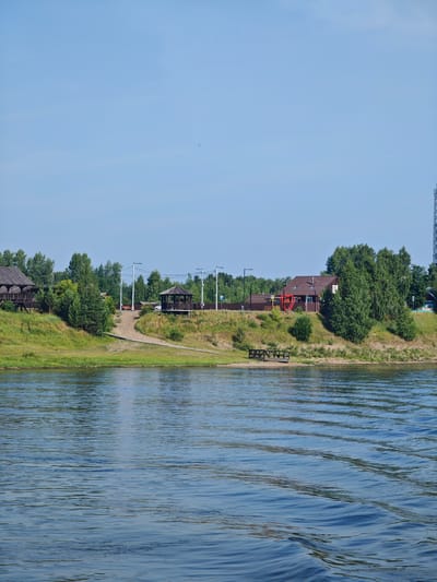 The image depicts a scenic view of a riverbank on a clear day. In the foreground, the blue water of the river displays gentle ripples, reflecting the sky and the surrounding greenery. The middle ground features a grassy embankment rising to a sandy area with a wooden path leading up to it. Several structures are visible on the embankment, including two wooden gazebos, one with a dark roof and lattice sides, and another with a lighter brown roof and open sides. A wooden house with a reddish-brown roof and white trim stands on the higher part of the embankment, adorned with a large red heart-shaped sculpture in front. Various trees, primarily deciduous and coniferous, are scattered along the embankment, creating a lush green backdrop. In the distance, a clear blue sky stretches across the horizon, with a few faint clouds. A tall radio tower is visible on the far right side of the image. The scene appears to be a tranquil riverside resort or recreational area, likely located in or near Galanino, Russia, given the context. There are no people visible in the image.