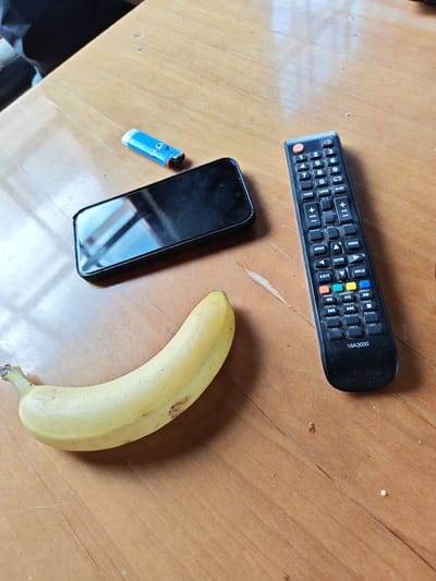 The image shows a wooden table with several objects on it. There is a yellow banana, a black smartphone with a reflective screen, a blue and white lighter with a cartoon face, and a black television remote control. The remote has numerous buttons, including number keys, volume and channel controls, playback buttons, and colored function keys. The remote also displays "16A3000" at the bottom. The lighting suggests an indoor setting, possibly a home, with light reflecting on the table and the phone screen. The background is out of focus but appears to be dark, possibly indicating furniture or a wall. There is no information about the location in Ичетовкины, Russia, as this is an unrelated detail.