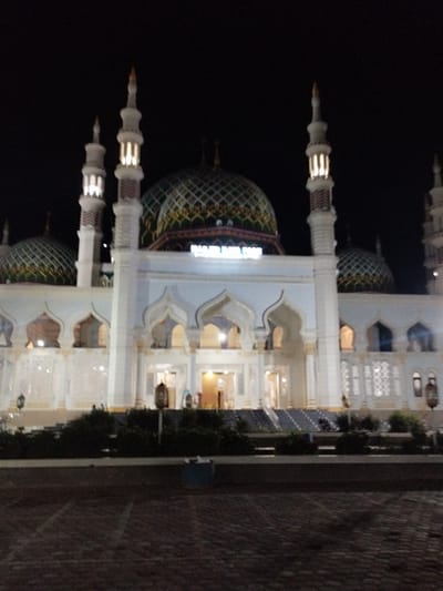 The image displays the Masjid Baiturrahim mosque at night in Kotapantonlabu, Indonesia. The mosque is brightly illuminated against a completely dark sky. The central structure features a large, ornate green and gold dome with a distinct geometric pattern, surmounted by an finial. Two prominent white minarets, each with a multi-tiered design and illuminated sections, flank the central dome. Several smaller green and gold domes are visible on either side of the main building. The mosque's facade is white, characterized by numerous pointed arches, intricate architectural details, and visible latticework or carved patterns. An illuminated sign above the central entrance clearly reads "MASJID BAITUR RAHIM". The foreground consists of a dark, paved surface, likely a courtyard or plaza. A low line of dark green shrubbery is situated in front of the mosque, with illuminated steps leading to its entrances. Several decorative lanterns or light fixtures are positioned at ground level along the facade. No individuals are discernible within the frame. A dark blue container is partially visible on the far right of the paved foreground.