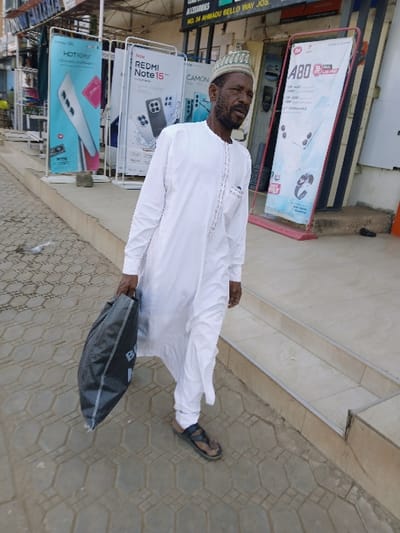 An adult male is depicted walking on a paved sidewalk in front of a commercial building. The man, with a beard and mustache, wears a white long-sleeved tunic (thobe) and a light-colored skullcap. He carries a dark grey fabric bag with white lettering (possibly "BALFOUR") in his right hand. His footwear consists of dark brown sandals. The sidewalk features grey hexagonal interlocking pavers. Behind the man, an elevated step leads into a building. The background reveals a shopfront with several vertical advertising banners for mobile phones and electronics, including "HOT 60i Pro+", "REDMI Note 15", "CAMON", and "ABO" branded items. Above the banners, a sign partially visible indicates "COMPUTERS ACCESSORIES NO. 34 AHMADU BELLO WAY JOS." This sign establishes the location as Ahmadu Bello Way, Jos. The image does not contain any visual information pertaining to Kungulu School, Zambia.