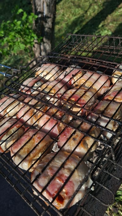 The image depicts a metal grill filled with food items. Specifically, it appears to be mushrooms wrapped in bacon, suggesting they are being cooked or grilled. The grill is made of a dark, possibly rusty metal, with a grid pattern that holds the food in place. The mushrooms are circular, light-colored, and wrapped with strips of bacon, which are pink and white with some browning from cooking. The background shows a glimpse of a green grassy area, indicating an outdoor setting. The image appears to have been taken in Сваткаўскі сельскі Савет, Belarus.