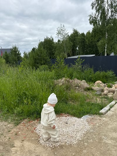 A young child, dressed in a white hooded tracksuit and a white hat, stands on a patch of white gravel in a construction area. The child is facing away from the camera, looking towards a pile of dirt and rocks. In the background, there is tall green grass, trees, and a dark fence. The sky is overcast.