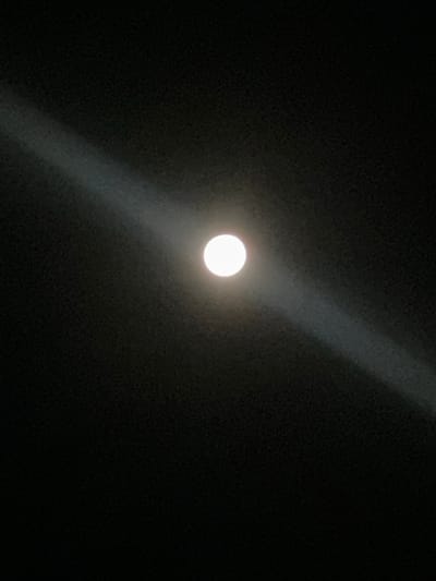 The image shows a full moon in a dark night sky. A faint band of light stretches diagonally across the sky, emanating from the moon. The moon is a bright white orb, slightly blurred, suggesting it was captured with a camera that may have had difficulty focusing on such a bright object against a dark background. The surrounding sky is uniformly black, indicating it is nighttime and there are no visible stars or clouds. The overall impression is of a serene, isolated lunar event. The location is specified as Sadovik, Bulgaria.