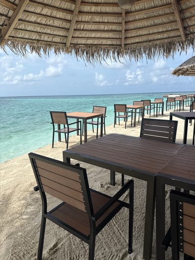 An outdoor dining area with tables and chairs is set up on a sandy beach next to a calm turquoise ocean under a thatched roof. In the foreground, a dark brown wooden table with a black metal frame is partially visible, with a matching chair in front of it. Behind this, several more tables and chairs are arranged on the sand, extending towards the water. The ocean is clear and shallow near the shore, transitioning to a deeper blue further out. The sky is partly cloudy. In the background, more thatched-roof structures are visible. The scene appears to be a restaurant or cafe located on a beach in Felidhoo, Maldives.