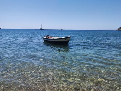 The image depicts a serene coastal scene in Zagrađe, Montenegro. In the foreground, a clear, shallow sea reveals a seabed covered with numerous smooth, multicolored pebbles. The water's surface is gently rippled, reflecting the bright, clear blue sky.
Further out in the water, a small, white and blue motorboat is moored. It has an outboard motor at the stern and a license plate that reads "BB-752". The boat is anchored by a rope leading out of frame to the right.
In the middle ground and background, the expanse of the Adriatic Sea continues, with several other vessels visible. To the left, a white catamaran with sails is anchored. Further out, a small, white speed boat is also present. There are a few buoys floating in the water, and some people can be seen swimming or wading in the distance. The horizon is a clean line where the sea meets the sky, with a hint of a hilly coastline on the far right. The overall atmosphere is one of calm and clear summer weather.