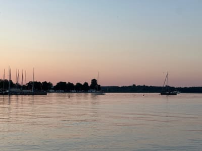 The image depicts a tranquil scene at a marina in Prien am Chiemsee, Germany, during twilight. The sky transitions from a pale blue at the top to a soft peach and pink hue near the horizon, indicating the sunset or sunrise. The calm water of the lake reflects the subtle colors of the sky and the silhouettes of various boats.
In the foreground, the rippling water is the dominant element, with gentle waves creating a textured surface that catches the fading light.
In the mid-ground, a marina is visible along the left side of the image, featuring a row of sailboats with their masts standing tall against the sky. Some of these sailboats appear to be docked. Further out on the water, a single white sailboat with a tall mast is sailing to the right, its hull dark against the water. To the right of this sailboat, a larger, dark-hulled boat, possibly a motor yacht, is also visible. A red buoy is present in the water to the left of the dark-hulled boat.
In the background, a treeline of dark green foliage forms a dense border along the far shore. Behind the marina on the left, a few white tents or awnings suggest a campground or event area. A single red buoy is positioned in the water in front of the treeline and to the left of the marina. The overall atmosphere is serene and peaceful, characteristic of a quiet evening by the lake. There are no visible people in the frame.