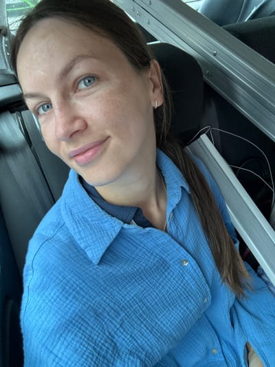 A woman with light skin, blue eyes, and brown hair pulled back, with a section draped over her left shoulder, is depicted in a medium close-up, high-angle portrait within a vehicle in Deinste, Germany. Her face shows visible freckles and a faint linear mark on her forehead. She is looking directly at the camera with a slight smile, revealing light pink lips. A small, silver-colored hoop earring is visible in her right ear. She wears a textured, light blue, long-sleeved button-up shirt over a darker blue collared garment, and a thin silver chain necklace is visible around her neck. A dark grey seatbelt crosses her left shoulder. In the background, to her right, multiple long, silver-colored metallic objects with visible rivets are positioned on a dark surface, potentially a rear seat, alongside a white cable. The visible interior of the vehicle is dark grey and black.
