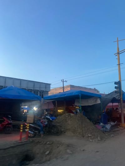 The image depicts a street scene in Keudelapang, Indonesia. The view is angled upward, showcasing a clear blue sky above buildings and street-level activity. Power lines stretch across the sky. Buildings are visible, some with blue tarps or awnings extending outward. A dirt or gravel embankment runs along the right side of the frame. Motorcycles are parked and possibly in use on the street. Red and yellow barricades are present. A pile of dirt or other material sits beside the street. The scene appears to be in a commercial or residential area with a mix of formal buildings and temporary structures.