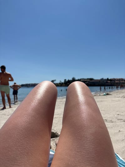 The image depicts a close-up view of a person's legs resting on a sandy beach. The legs are tanned and appear to have sunscreen or lotion applied, as evidenced by the subtle shimmer on the skin. In the background, a body of calm water, likely the Atlantic Ocean, is visible, with several people wading or standing in the shallow water. Further in the distance, a coastline with buildings and trees can be observed. The sky is a clear, bright blue. The scene suggests a sunny day at a beach in the city of Pontevedra, Spain, a popular coastal region known for its beaches and natural beauty.