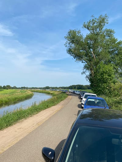 The image depicts a traffic jam of cars on a paved road next to a calm, narrow body of water. The road curves to the left, and a long line of vehicles stretches into the distance. The water is bordered by green grass and reeds. In the background, more cars are visible, and beyond that, a line of trees and some buildings. The sky is bright blue with a few scattered white clouds. The scene appears to be in a rural or semi-rural area, possibly on the outskirts of Meppel, Netherlands, given the landscape and the presence of a canal-like waterway. The time of day is likely daytime, given the bright lighting and clear sky.