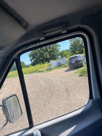 The image is taken from inside a vehicle, looking out through the driver's side window. The foreground shows the interior of the vehicle, including the grey headliner, a sun visor, and part of the window frame and dashboard. A side mirror is visible on the left.

Outside the window, a gravelly area extends into the midground. In the distance, a black Infiniti FX35 SUV is parked on the gravel. The SUV has a license plate that reads "A PR 9508". Behind the SUV, there is a light-colored concrete structure with a fountain-like element.

The background features a grassy field with scattered yellow wildflowers. Large, mature green trees are prominent on the left and in the background, suggesting a rural or semi-rural setting. The sky is clear and blue. The scene appears to be in the vicinity of Poroy, Bulgaria, given the information provided. There are no people visible in the image. The time of day appears to be daylight, with bright sunlight casting shadows.
