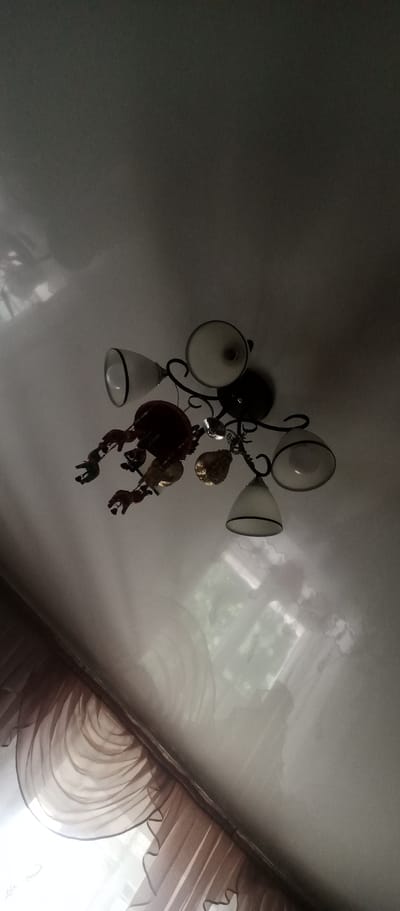 This image shows a chandelier with four lamps hanging from a light-colored ceiling. The chandelier has a black metal frame with curved arms. Three of the lamps have white glass shades, while one has a clear glass shade. Attached to the chandelier are several decorative elements, including a reddish-brown object that resembles a lobster or crab, and several clear or golden-colored crystal-like pieces. One of the lamps appears to have a lightbulb inside. The chandelier is positioned above a window with sheer, ruffled brown curtains. The ceiling appears to be a reflective surface, showing a distorted reflection of the chandelier and possibly the window. The overall lighting suggests it is daytime, with light coming through the window, creating shadows on the ceiling. The scene appears to be indoors, likely a room in a home. There is no visible text in the image.