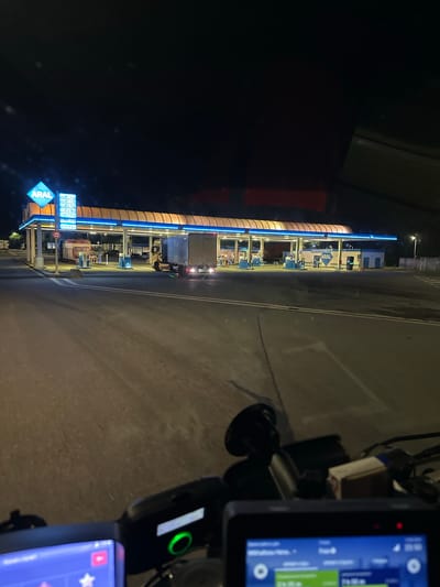 The image shows a night scene at an ARAL gas station, identified as being located in Luckau, Germany. The gas station is illuminated with bright lights, highlighting the canopy, pumps, and the ARAL logo. A truck is parked at one of the pumps. In the foreground, a dashboard with multiple screens and devices is visible, suggesting the image was taken from inside a vehicle. The overall environment appears to be an open area, likely a highway rest stop or a service area, with asphalt ground and some vegetation in the background.