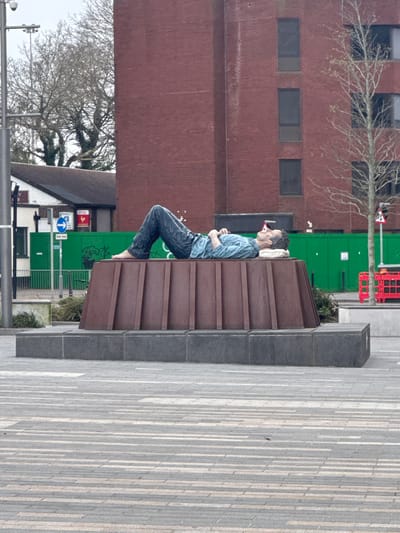 The image displays the public sculpture "The Sleeping Man" by artist Sean Henry, situated in Jubilee Square, Woking, United Kingdom. The sculpture depicts a life-sized male figure lying supine, barefoot, wearing blue jeans and a light blue long-sleeved collared shirt. His hands are clasped over his chest, and his head rests on a white, cushion-like object. The figure is mounted on a dark brown, multi-faceted metal pedestal, which in turn sits on a dark grey rectangular plinth. The immediate foreground is a pedestrian area paved with alternating light and dark grey rectangular blocks arranged in a linear pattern.
In the background, a tall, multi-story building constructed of red brick with vertically stacked, dark-framed rectangular windows is prominent. To the left, a smaller, white-painted building with a dark roof is partially visible, bearing the lettering "Y B CLUB" on its facade. Green construction hoardings, exhibiting black graffiti, are positioned behind the sculpture and to the right of the white building. A blue "One Way" traffic sign is affixed to a pole near these hoardings. Further to the right, red temporary barriers are present. Several deciduous trees, mostly bare of foliage, are visible against the overcast sky, with a younger tree positioned to the right of the sculpture. A grey utility pole equipped with a CCTV camera is on the far left of the scene.