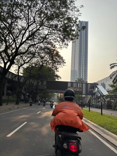 In Kelapa Dua, Indonesia, a person wearing an orange jacket with a Shopee logo and a black helmet rides a black scooter on a city street. The scooter is in the foreground, with its rear taillight illuminated. In front of the scooter, several other scooters are visible on the road, moving in the same direction. The street is paved and marked with white lines. To the left of the road, a large tree with dense branches is silhouetted against the sky, and a pedestrian bridge spans across the road. Further down the street, a tall, modern skyscraper with a light-colored facade stands against a hazy sky. Palm trees and other greenery are present on the right side of the road, along with a building and a sidewalk. The scene appears to be from a mid-morning or late afternoon, with the sun casting long shadows and creating a warm, diffused light.