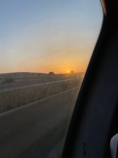 A sun is setting on a rural landscape, casting an orange glow across the sky. The sun is a bright orb near the horizon, partially obscured by a solitary tree. Fields stretch out towards rolling hills in the distance, with a road or highway running through the middle ground. A dark, possibly car interior, element is in the foreground, obscuring the left side of the scene. The sky transitions from a pale blue at the top to a warm orange and yellow near the horizon. The image appears to be taken from a moving vehicle. The location is Ouled Djilali, Algeria.