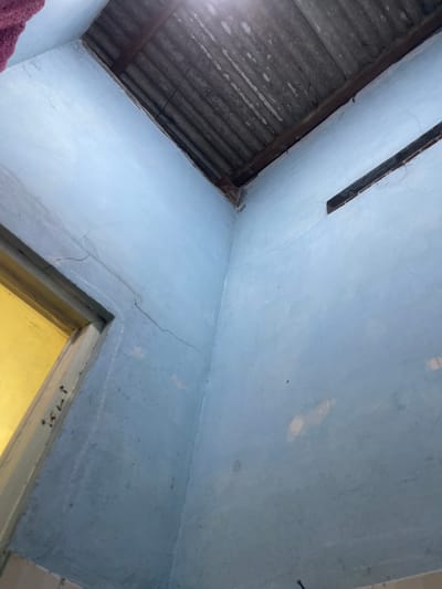 A low-angle, wide shot captures an interior corner of a room with light blue walls and a corrugated tin roof. The room appears unfinished or in disrepair, with visible cracks in the plaster of the walls and exposed wooden beams supporting the roof. A doorway with a yellow interior is visible on the left, leading out of the frame. The scene is devoid of people or any clear indication of recent activity, suggesting an abandoned or unused space. The lighting is dim, with light filtering from the opening in the roof and through the doorway. The overall impression is one of a simple, rural dwelling in a state of neglect. The context places this scene in Anggaswangi, Indonesia, a location known for its rural landscapes and traditional housing styles.