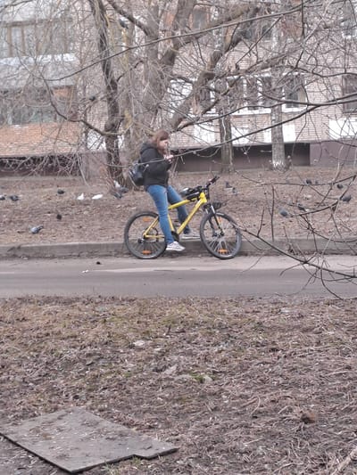 An outdoor scene in Yegoryevsk, Russia, depicts a female individual paused on a paved road, straddling a bright yellow mountain bike. She is positioned centrally, wearing a dark padded jacket, blue jeans, and light-colored athletic shoes, with a dark backpack over her left shoulder. She holds a small, light-colored electronic device, looking down at it. To the left, beyond a concrete curb, an area of dry, sparse vegetation and fallen leaves is populated by approximately two dozen pigeons, primarily grey with some white individuals. Numerous bare deciduous trees with intricate branch networks are present, partially obscuring multi-story brick buildings in the background, one with visible white-painted sections. The immediate foreground features additional dry ground cover and a rectangular concrete slab. The scene is illuminated by overcast light, consistent with late autumn or early spring conditions.